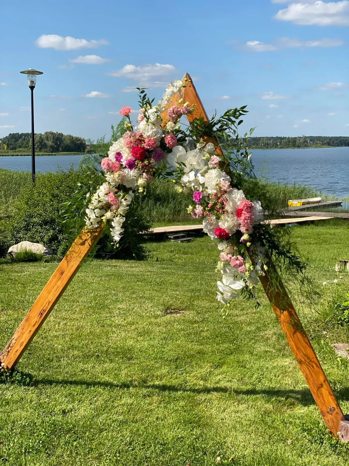 Drewniana brama ślubna w kształcie trójkąta, przybrana różowymi i białymi kwiatami, na trawniku nad jeziorem