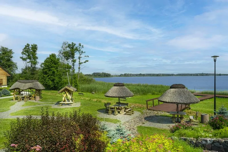 Ogród nad jeziorem — altany kryte strzechą, ławy i rabaty, w tle tafla wody