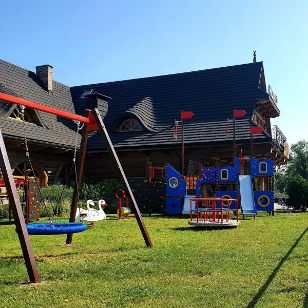 Plac zabaw z drewnianą wieżą i zjeżdżalnią, huśtawki na trawniku przy budynku