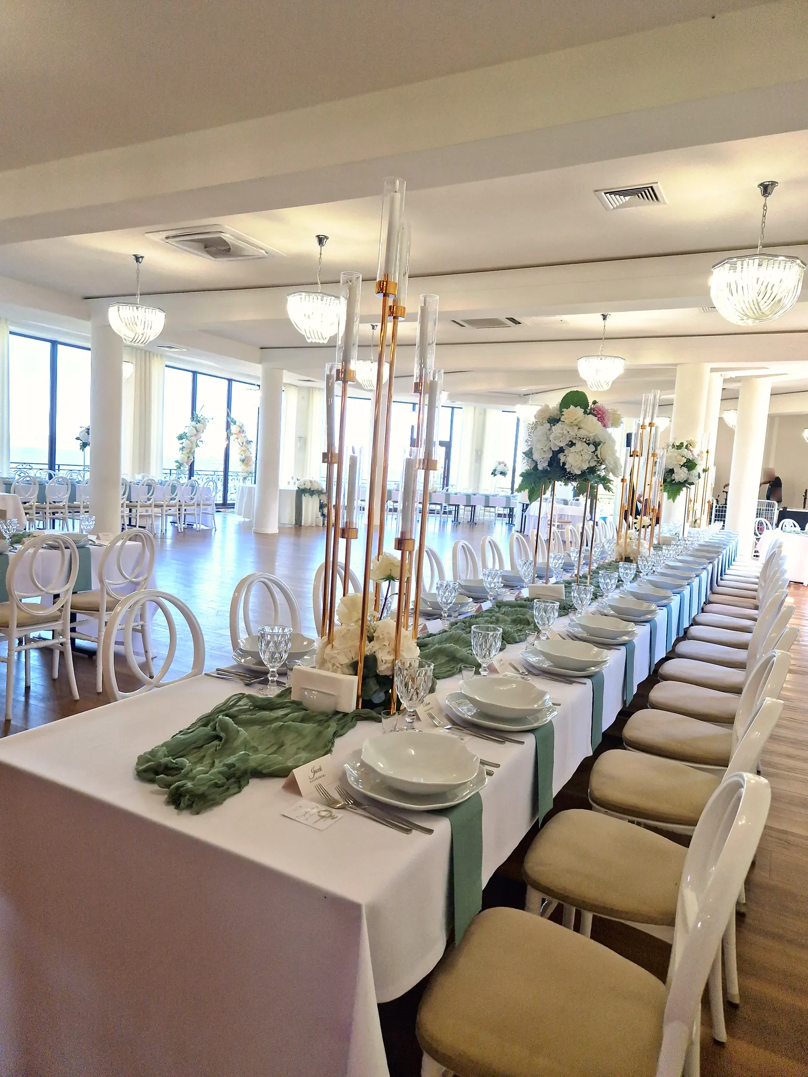 Sala Złota nakryta do wesela — długie stoły wzdłuż całej sali, złote świeczniki z kwiatami, białe słupy i panoramiczne okna na Jezioro Łabędź