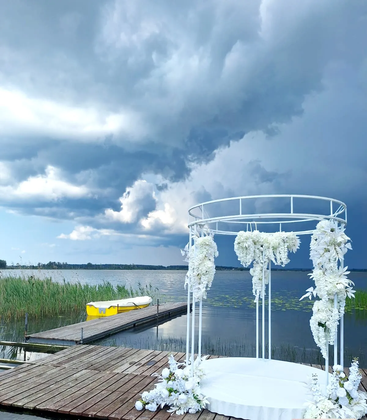 Okrągła biała brama ślubna z kaskadami białych kwiatów, ustawiona na drewnianym pomoście nad jeziorem, nad wodą burzowe chmury, przy pomoście zacumowana żółta łódka